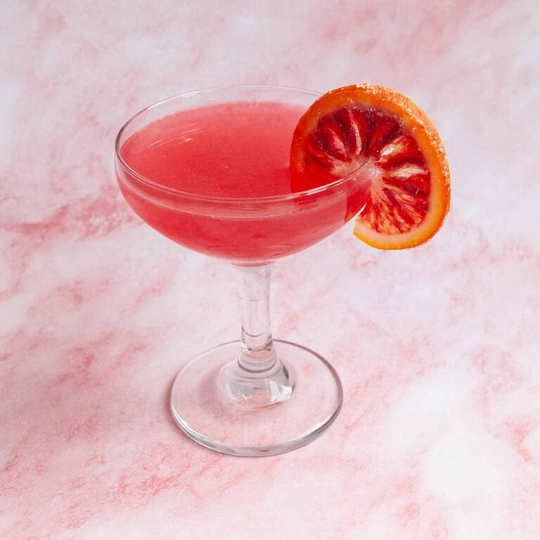 A pink cocktail in a coupe glass with an orange wheel on the side.