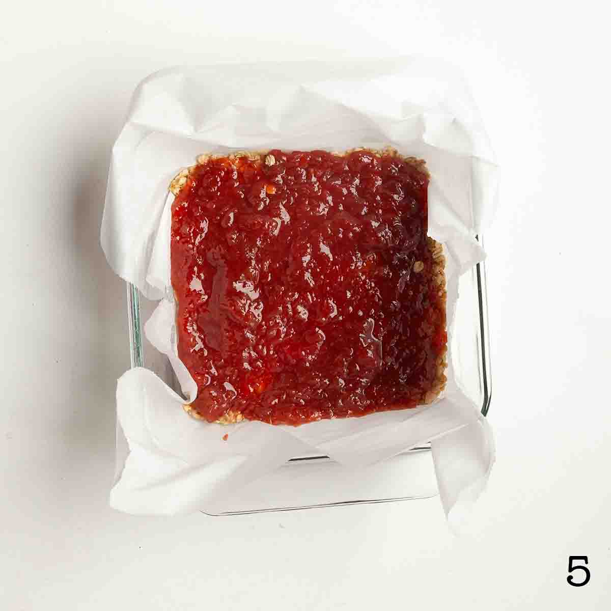 A layer of jam on top of an oat bar base in a square baking dish.