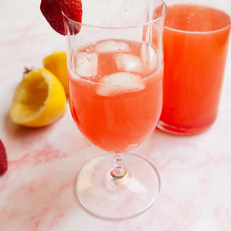 A glass of strawberry mint lemonade with ice and a pitcher in the background.