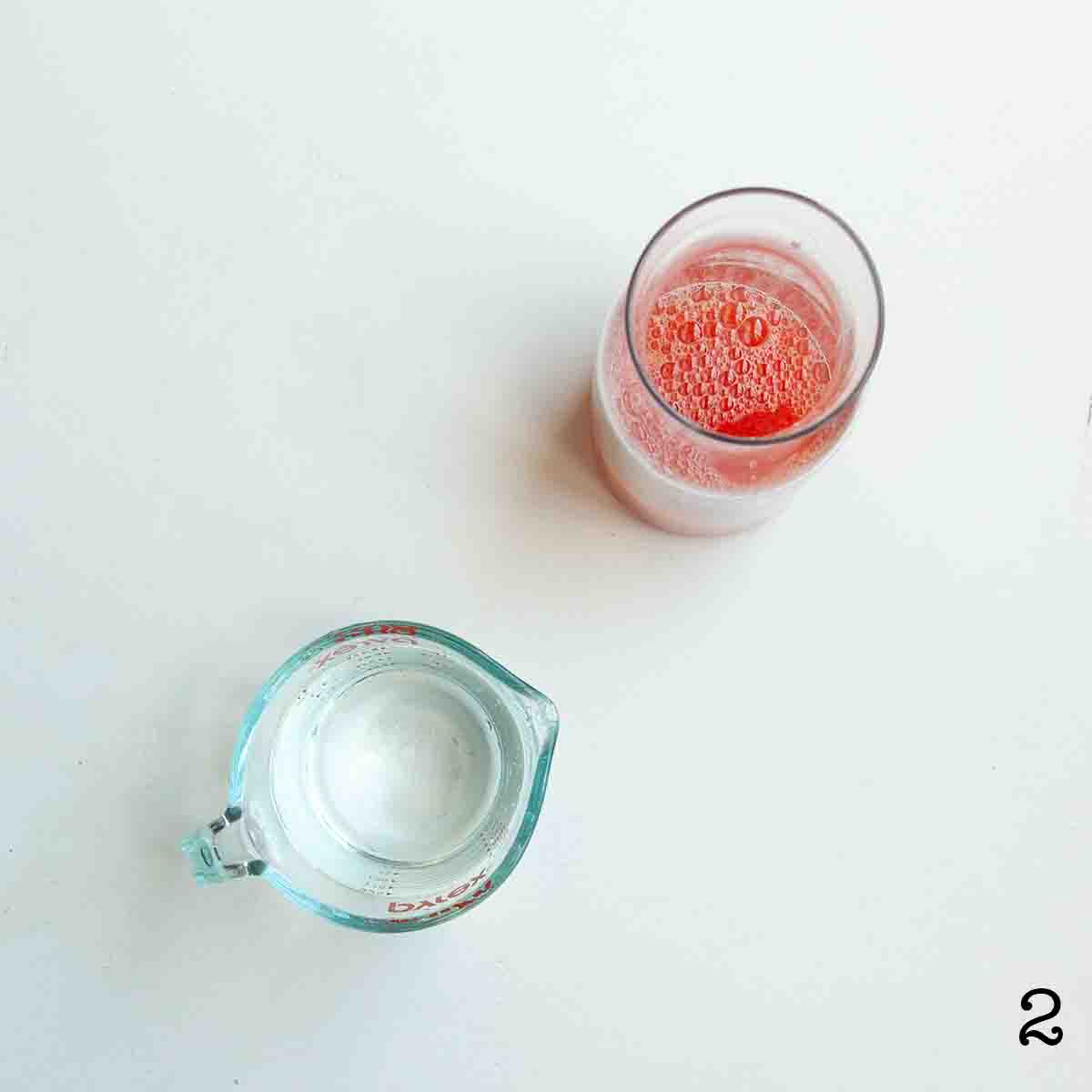 A pitcher of strawberry lemonade with a measuring cup of water on the side.