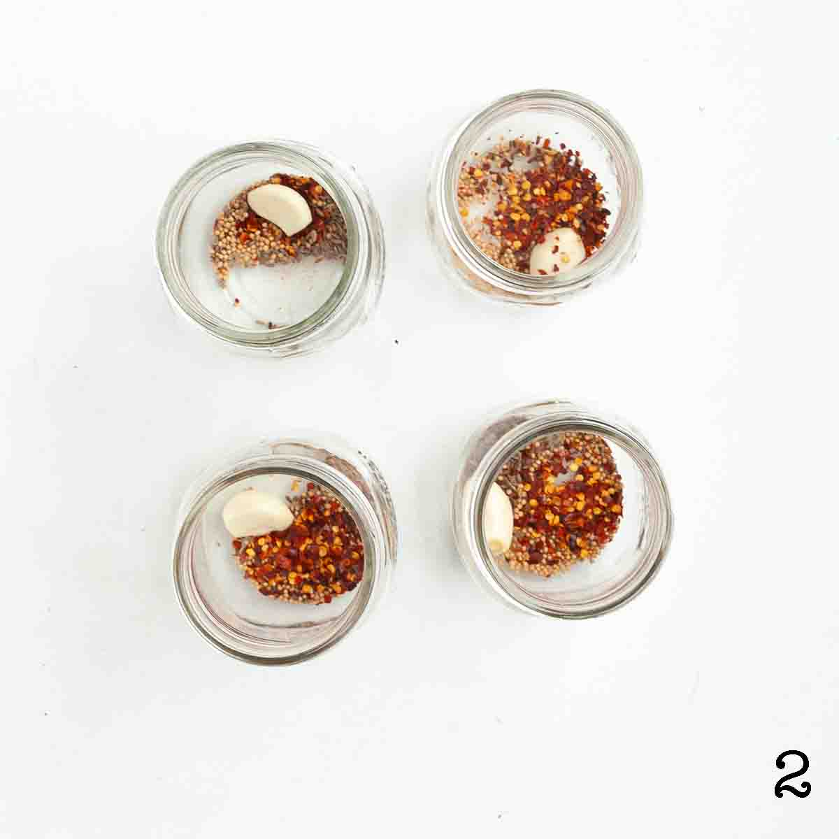 Four jars each with a garlic clove, pepper flakes, mustard seed, and dill seed.