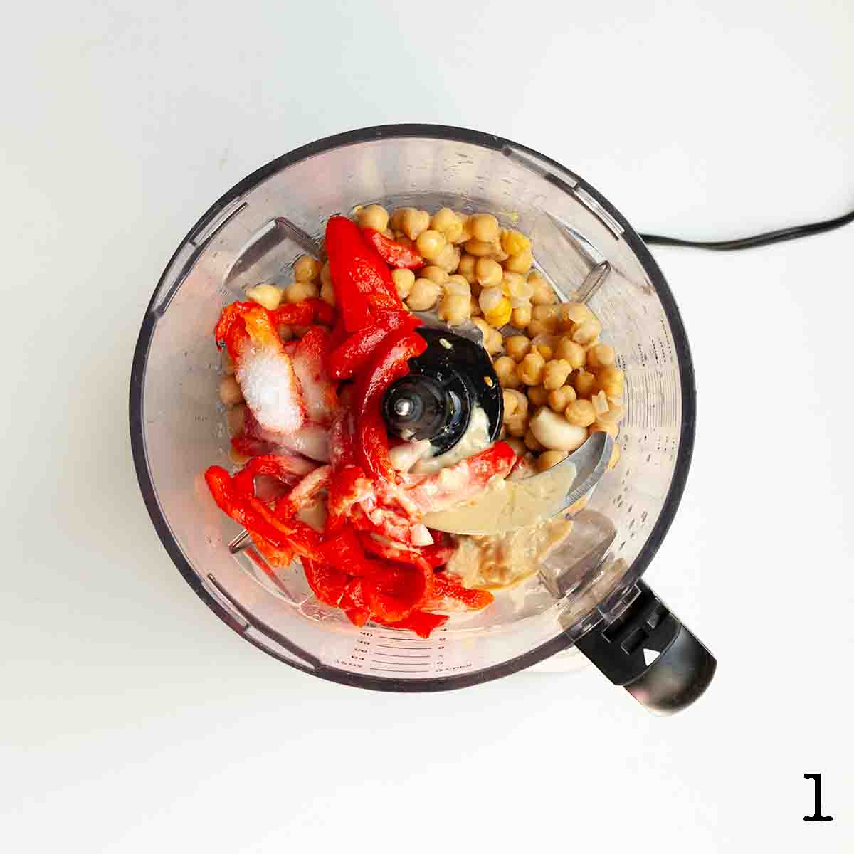 A food processor with roasted red peppers, chick peas, salt, and tahini in it.